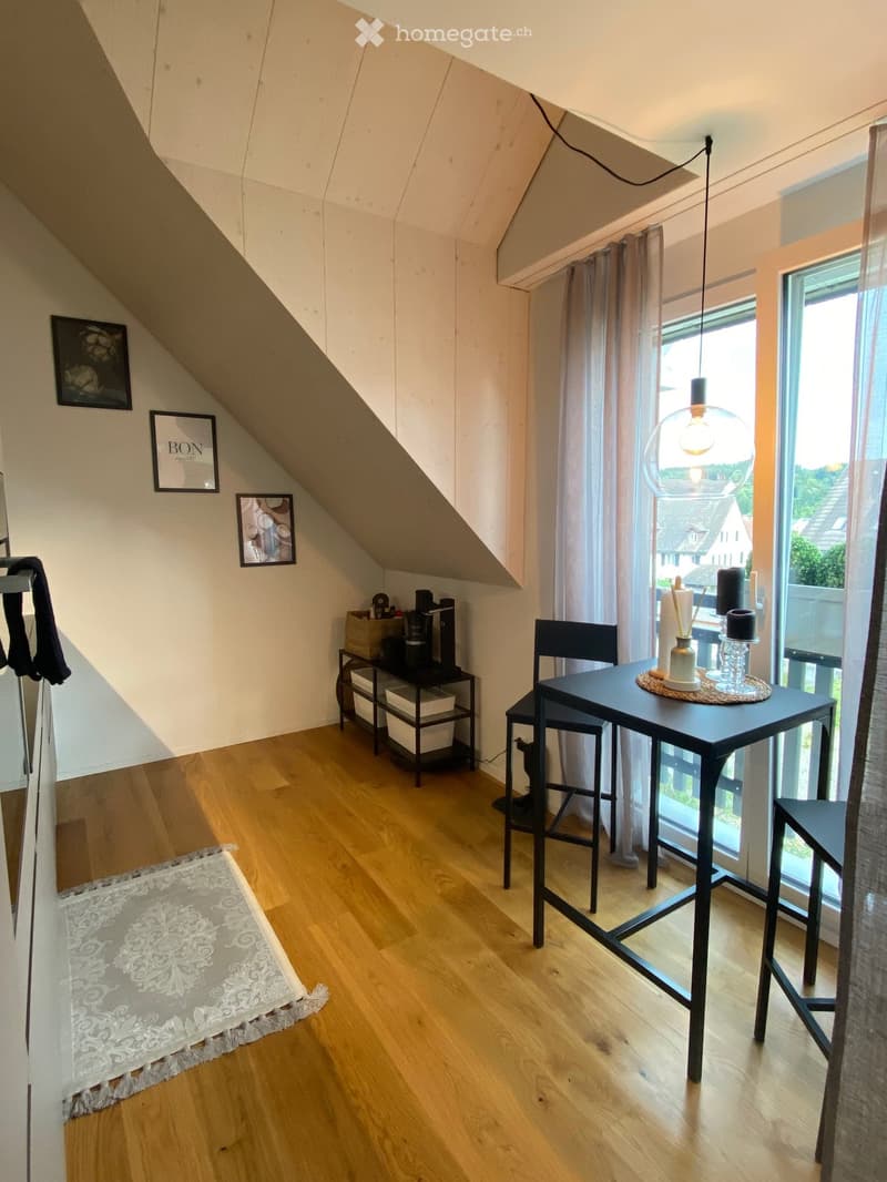 A cozy attic room with wooden flooring, large windows, and modern decor including a table, chairs, and a hanging lamp.