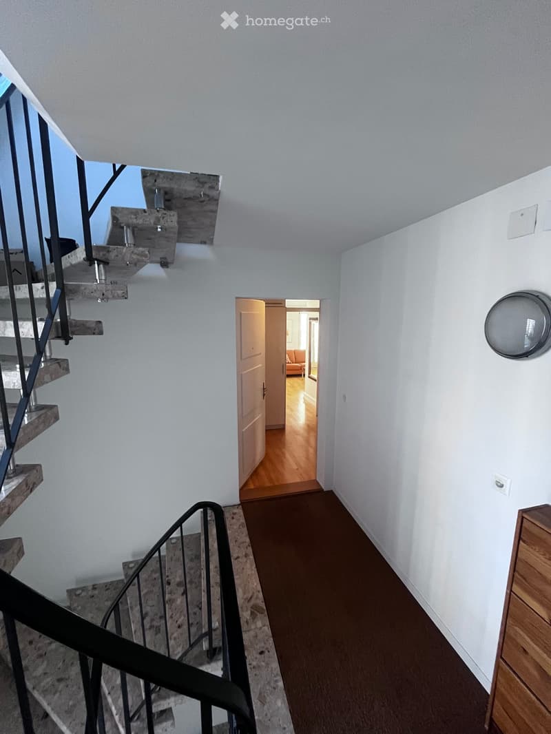 A modern hallway with a wooden floor, white walls, and a staircase leading upwards. A round wall-mounted light fixture is visible on the right side, and a partially open door leads to another room with a visible sofa.
