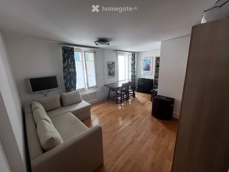 A modern living room with a light-colored sectional sofa, a wooden dining table with chairs, a flat-screen TV mounted on the wall, large windows with curtains, and a radiator below one window. The floor is made of light wooden planks.