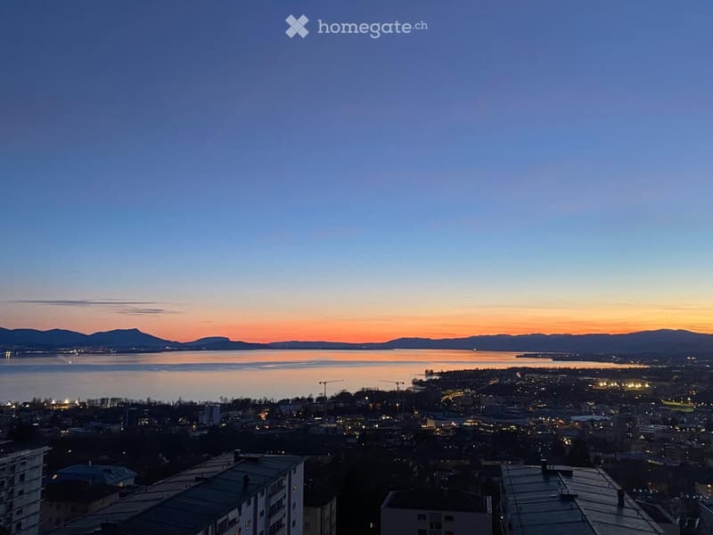 Panoramic view of a cityscape at sunset, featuring a body of water, distant mountains, and illuminated buildings.