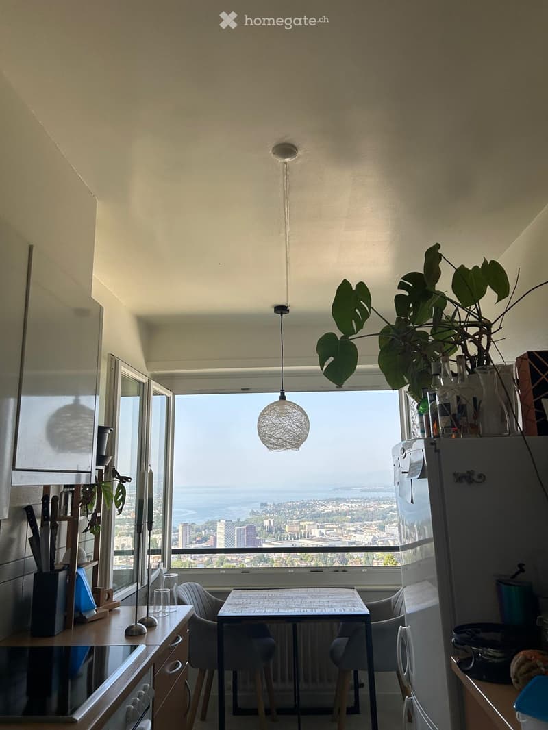 Modern kitchen with a view of the city and sea, featuring a refrigerator, dining table, chairs, pendant light, knife block, candle, and plant.