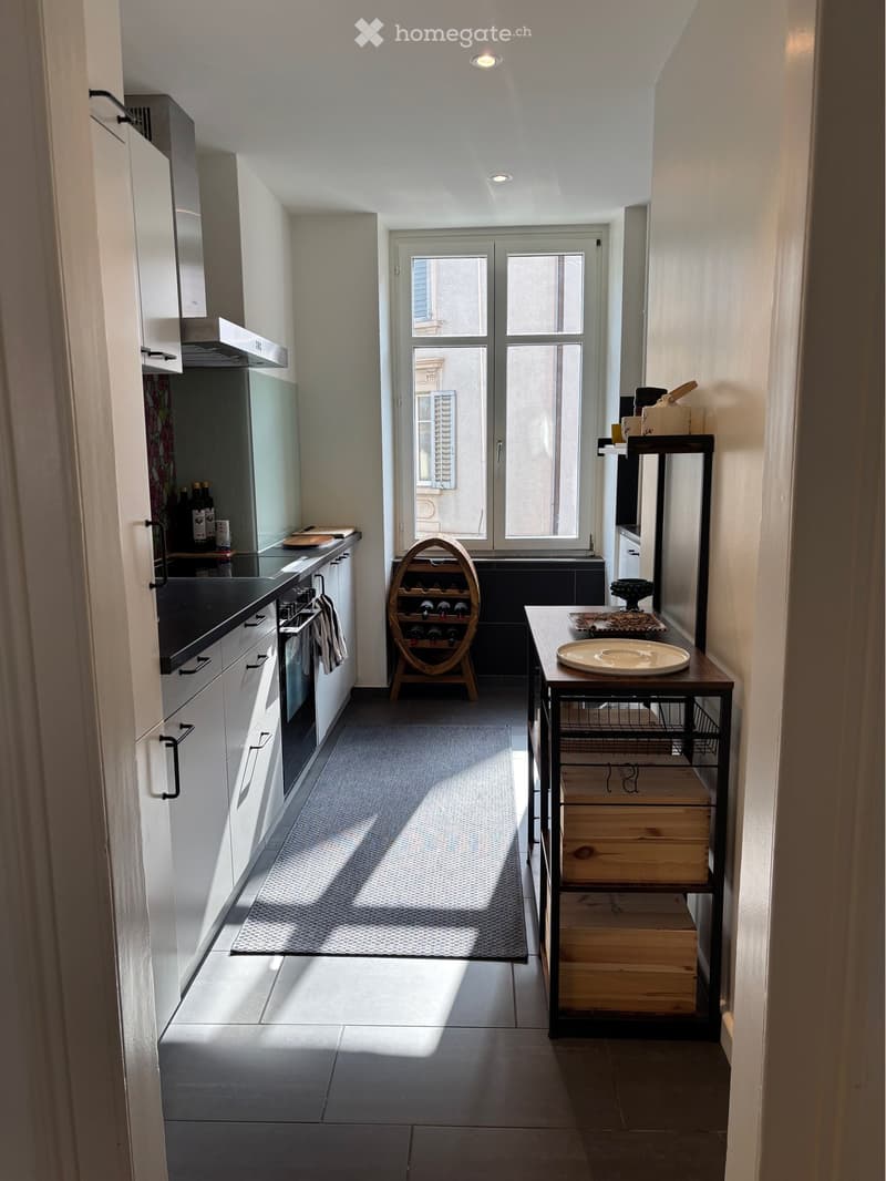 Modern kitchen with white cabinets, stainless steel appliances, a wine rack, a small table with a plate, and a shelf with wooden crates.