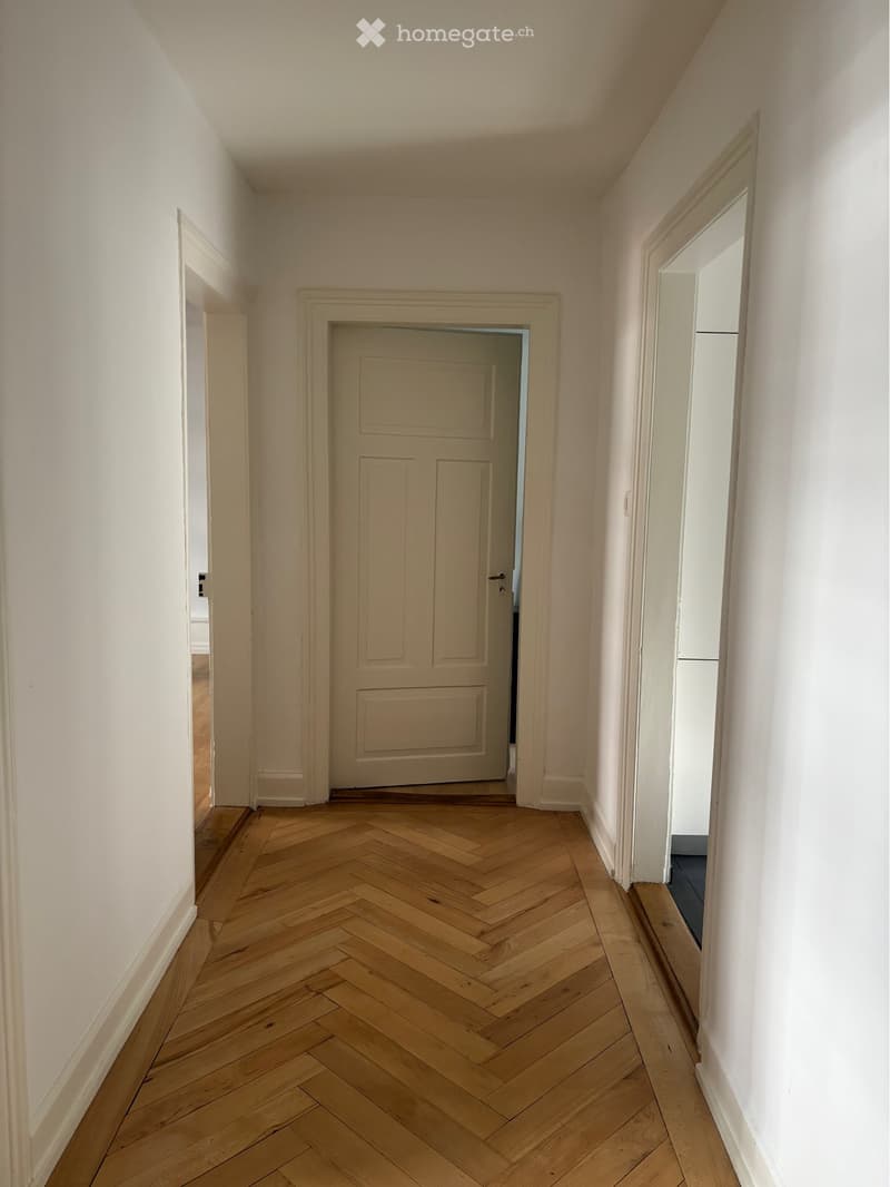 Empty hallway with herringbone wooden flooring, white walls, and a closed door at the end.