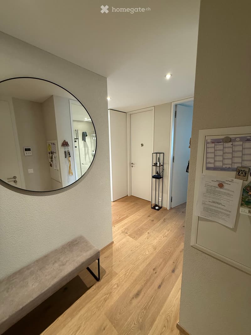 A well-lit hallway with wooden flooring, a small bench, a round mirror, and a small black shelf with items. Doors lead to other rooms, and there are notices and a calendar on the wall.