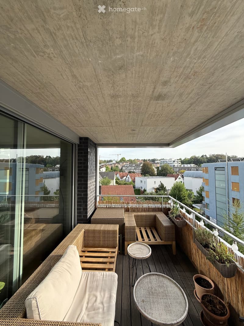 Modern rooftop terrace with wooden decking, wicker sofa, round tables, and potted plants, overlooking a cityscape.