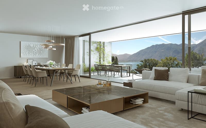 The image shows a modern and spacious open-plan living and dining area with large windows offering a stunning view of the mountains and lake. The living room features a comfortable white sofa and a low, rectangular coffee table with storage compartments.