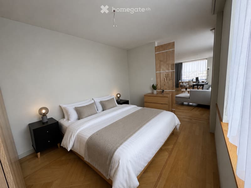 Modern bedroom with a large bed, nightstands, lamps, and a small living area with a sofa and table.