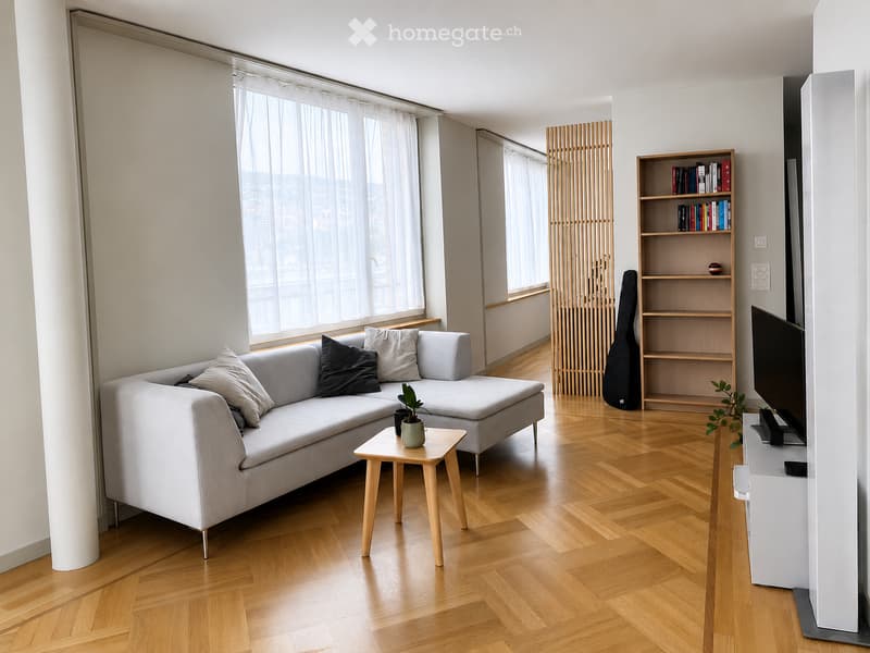 Modern living room with light wooden flooring, large windows with sheer curtains, grey sectional sofa with throw pillows, small wooden coffee table, bookshelf, guitar case, and TV stand.