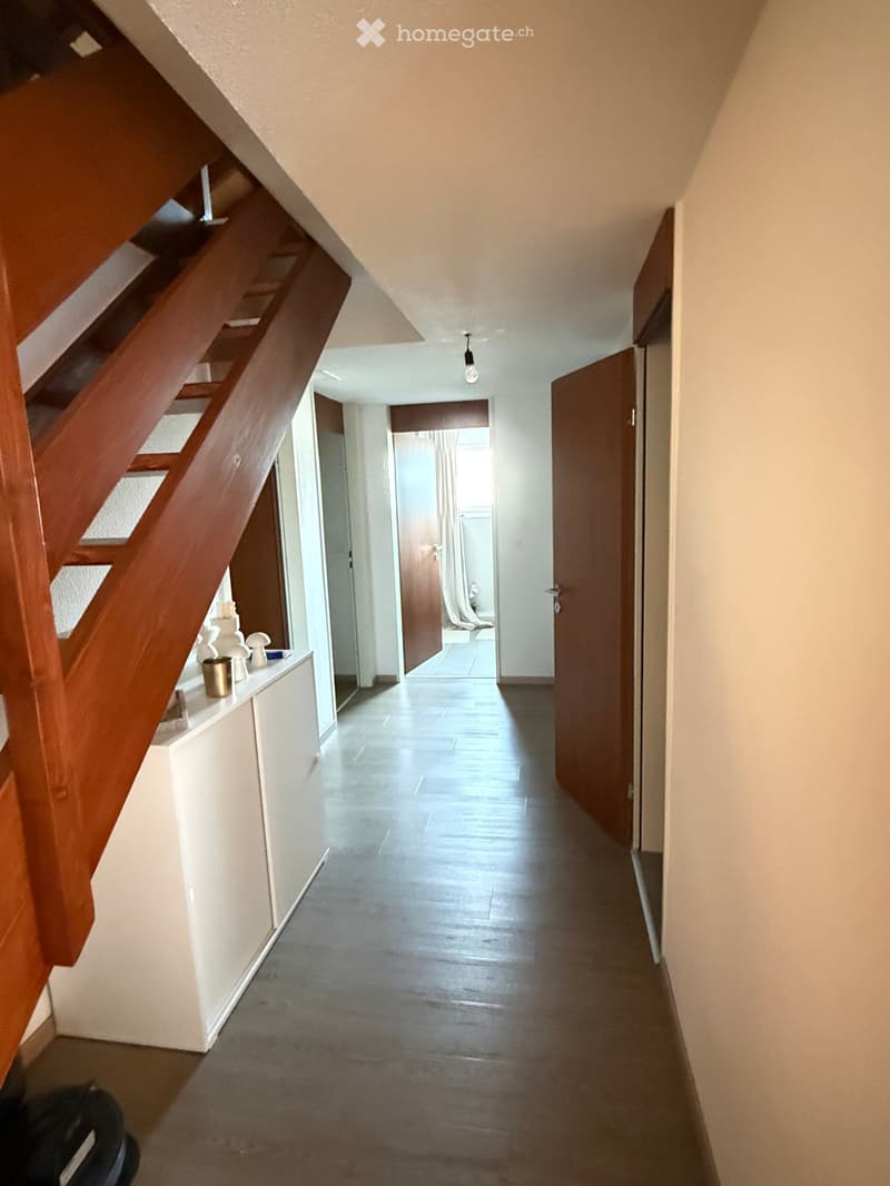 A modern hallway with wooden staircase, grey tiled floor, white walls, and several doors leading to rooms.