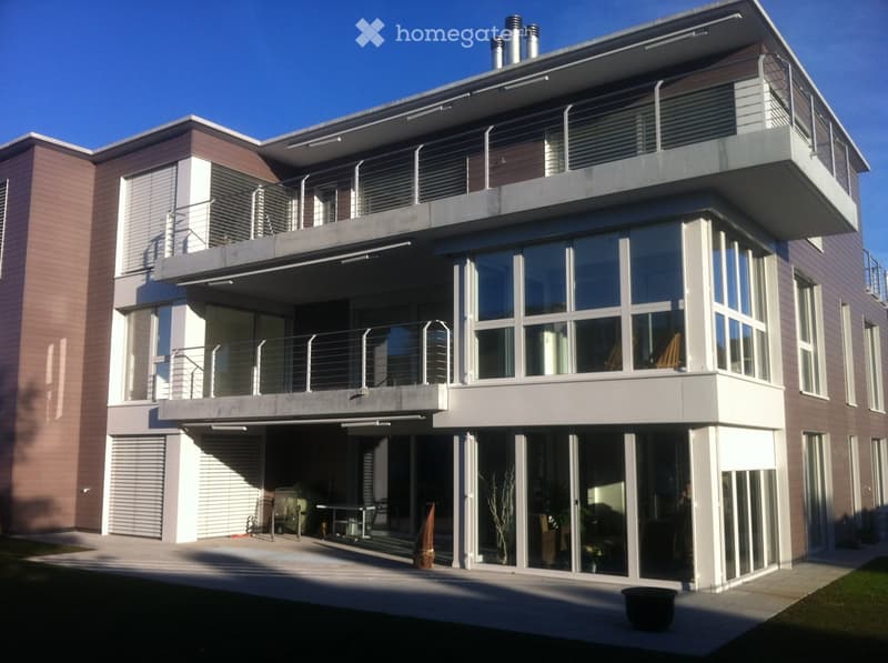Modern two-story house with large windows, balconies, and a covered entrance area. Features include a raised ground floor, multiple balconies, and a contemporary design with a mix of brick and concrete finishes.