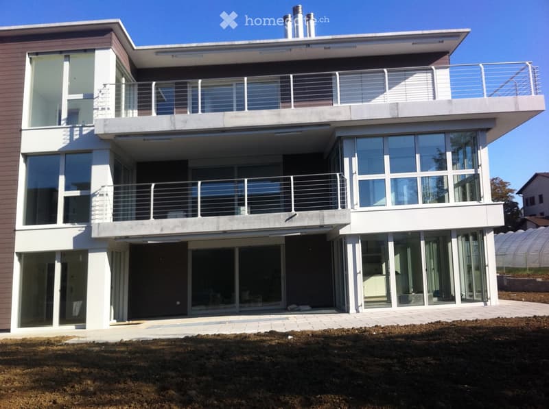 Modern two-story house with large glass windows, balconies, and a raised ground floor. The exterior features a combination of white and brown tones with metal railings on the balconies.