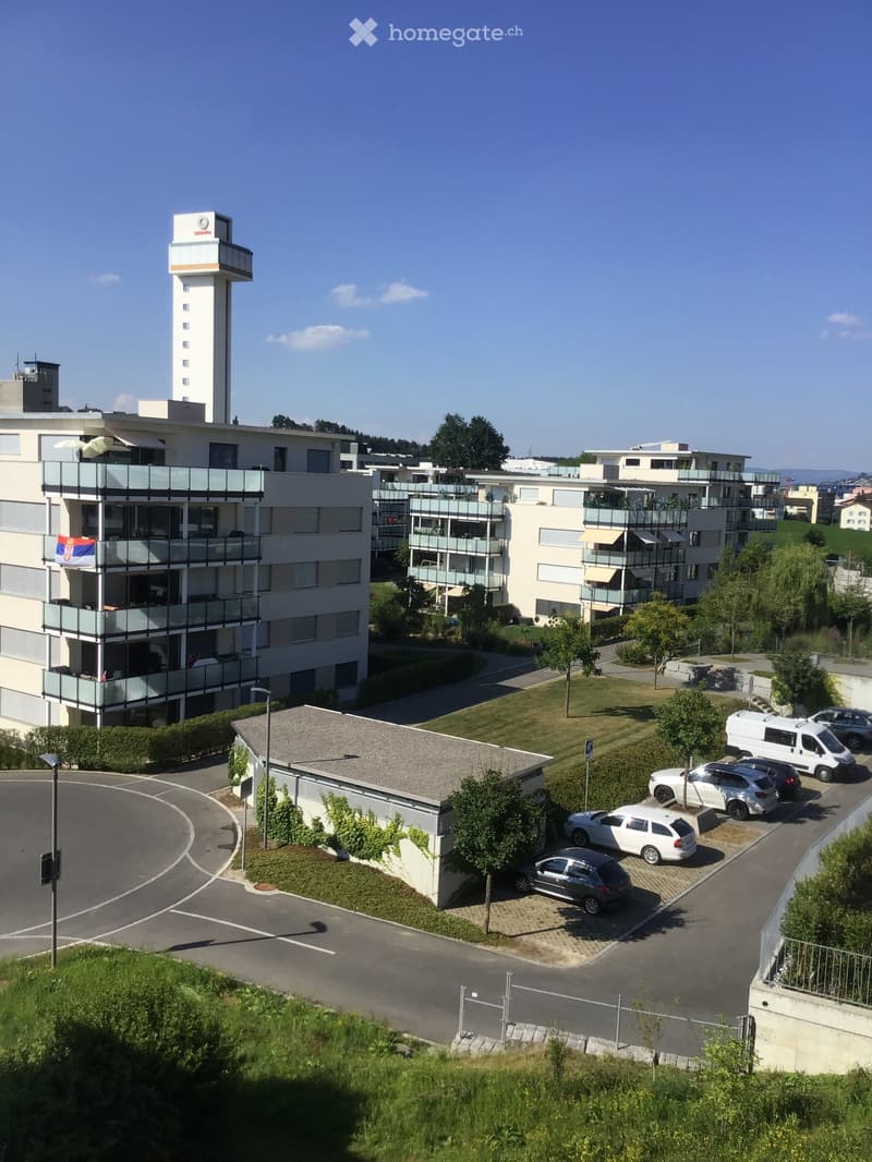 Modern multi-story residential complex with balconies, parking area, and surrounding greenery.