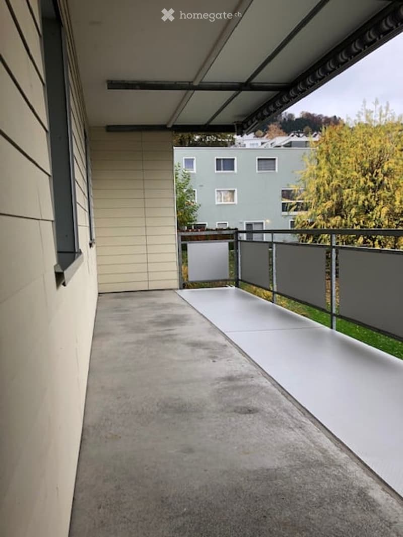 Modern balcony with concrete floor, metal railing, and a covered section. The view shows a neighboring building and greenery.