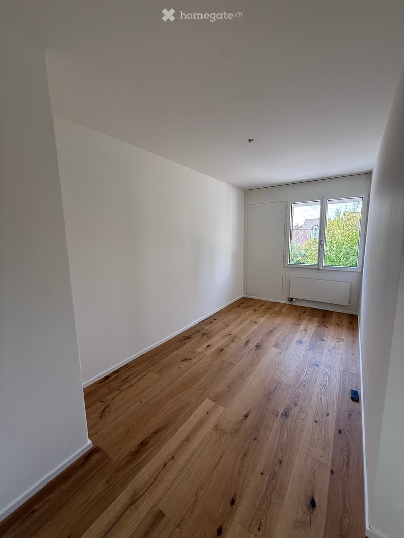 Empty room with light wooden flooring and white walls, featuring a window with a view of greenery and a radiator below it.