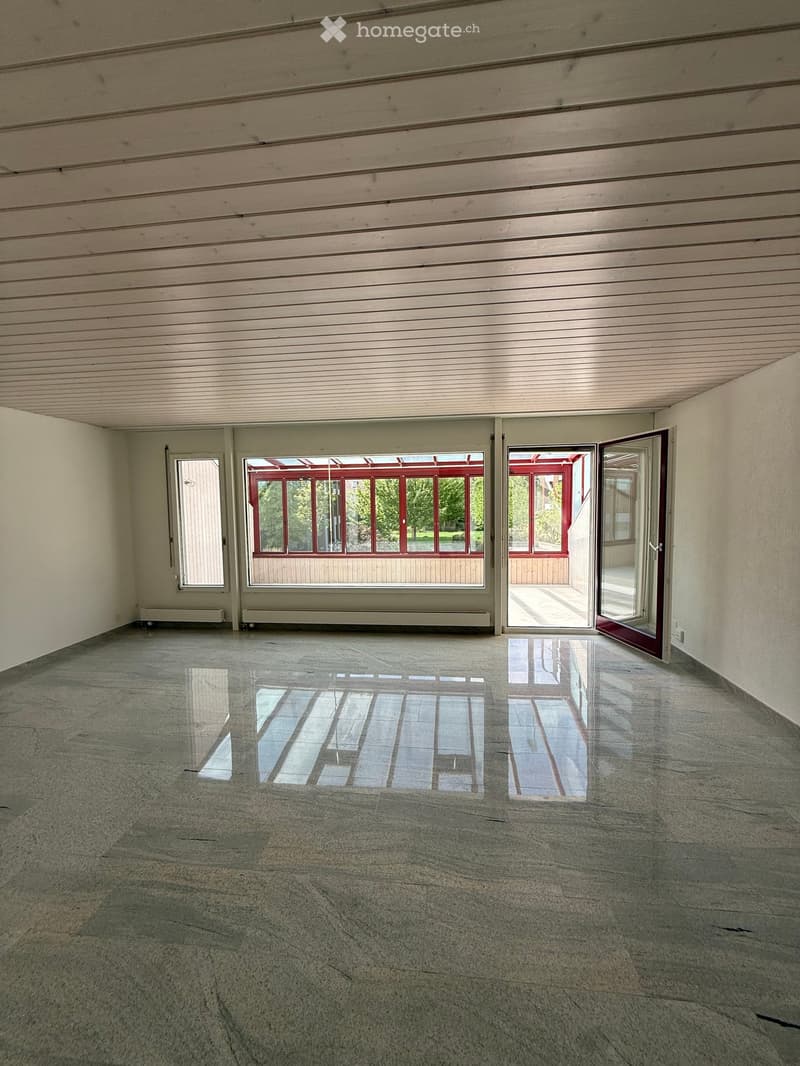 Empty room with polished concrete floor, white walls, and large windows with red frames. The space is well-lit with natural light reflecting off the shiny floor.