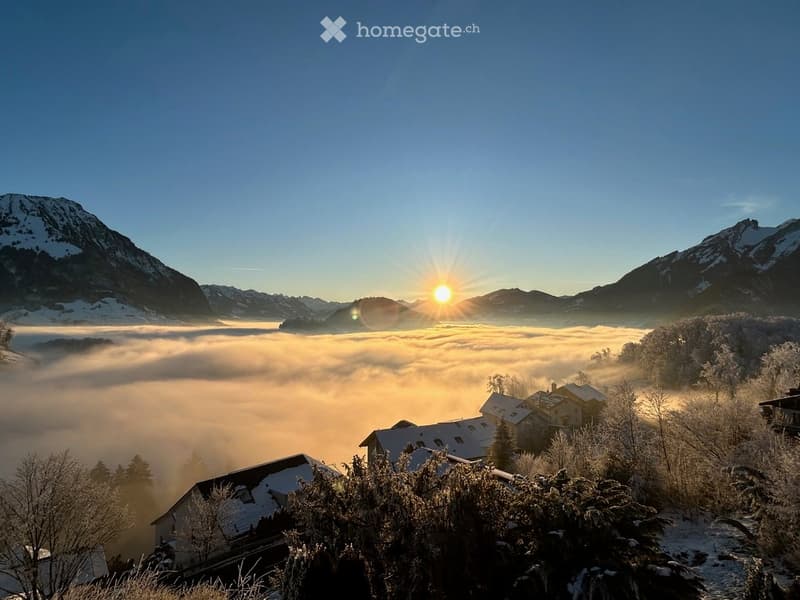 A serene mountain landscape at sunrise, with snow-covered peaks, a valley filled with mist, and a few houses nestled among trees.