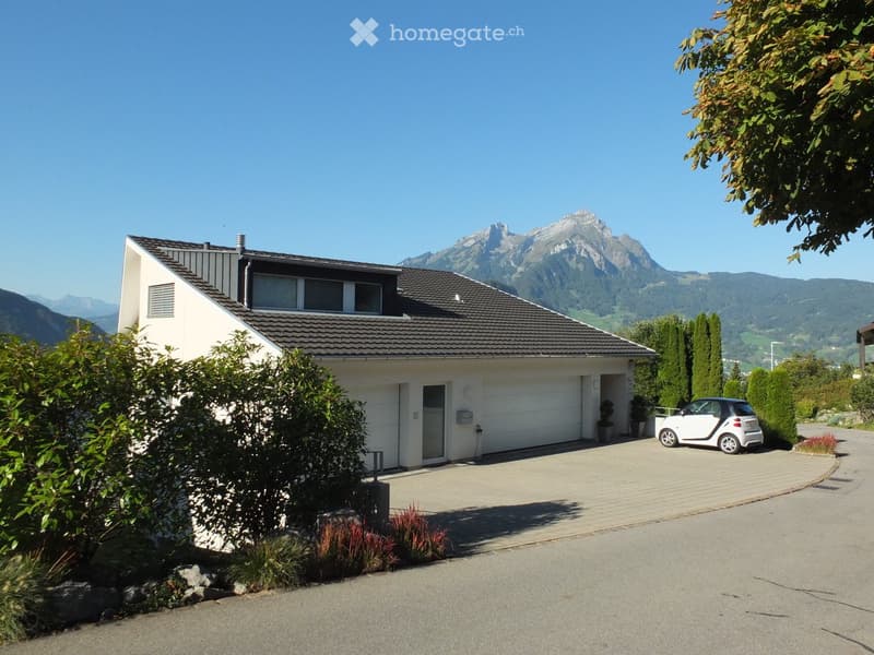 Modern two-story house with a grey roof, white walls, garage, and driveway, surrounded by greenery and a scenic mountain view.