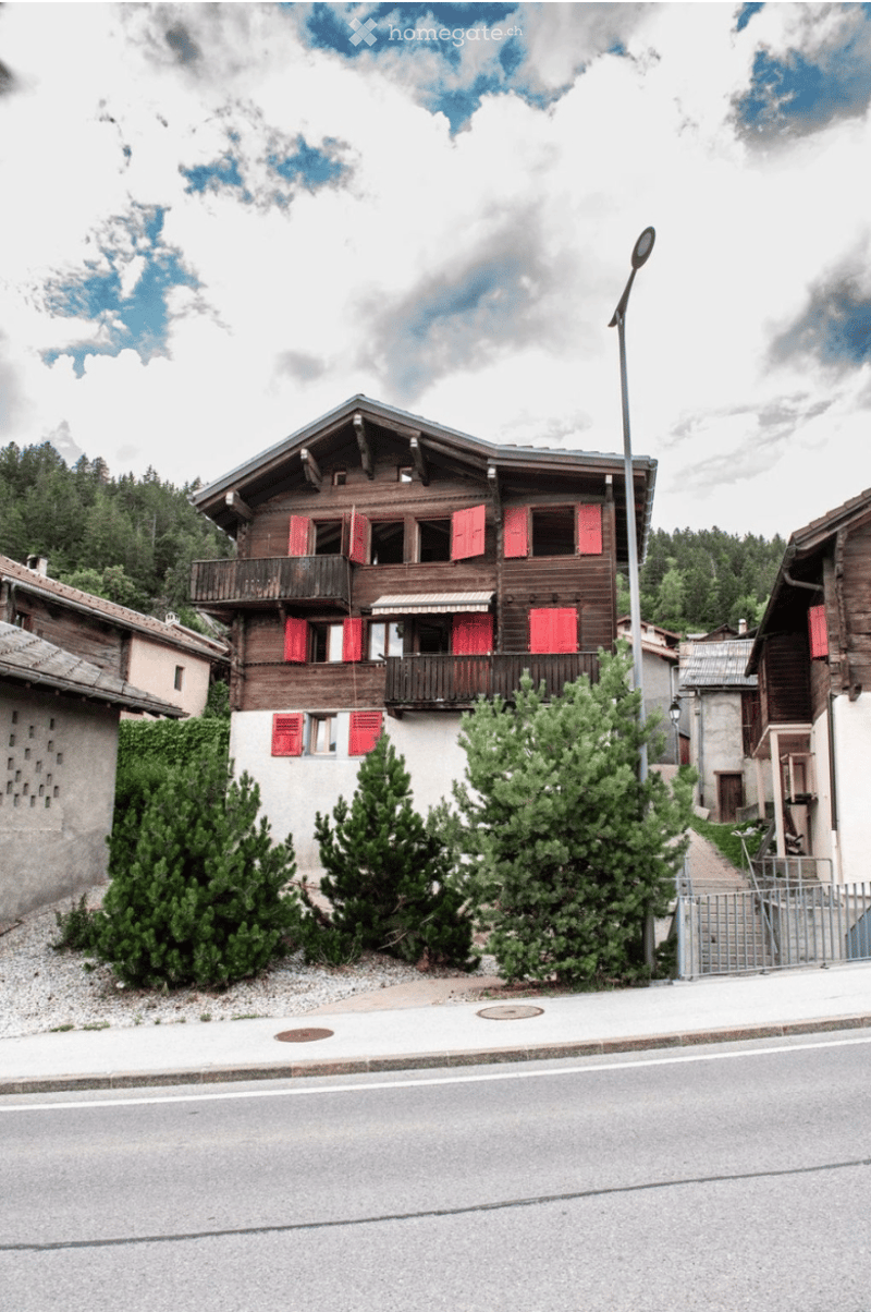 Traditional wooden chalet with red shutters, balconies, and surrounding greenery in a village setting.