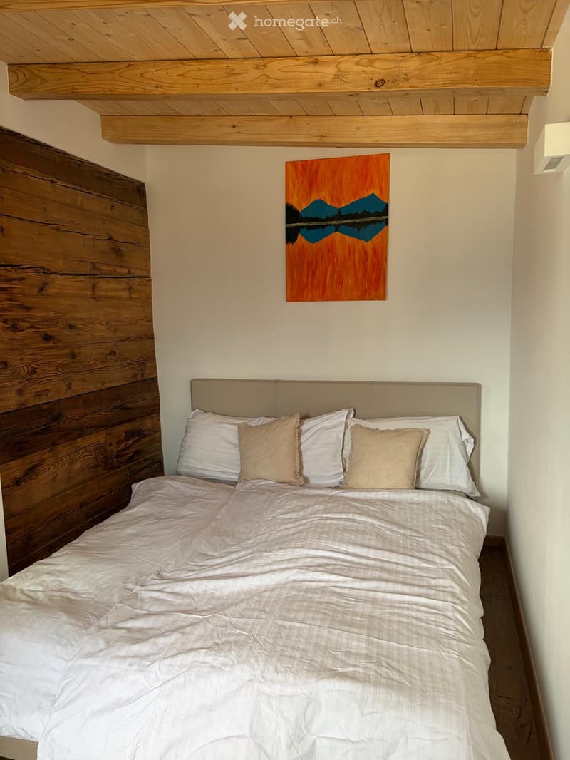A cozy bedroom with a neatly made bed, white bedding, and a wooden headboard. A painting of a mountain landscape hangs on the wall above the bed.