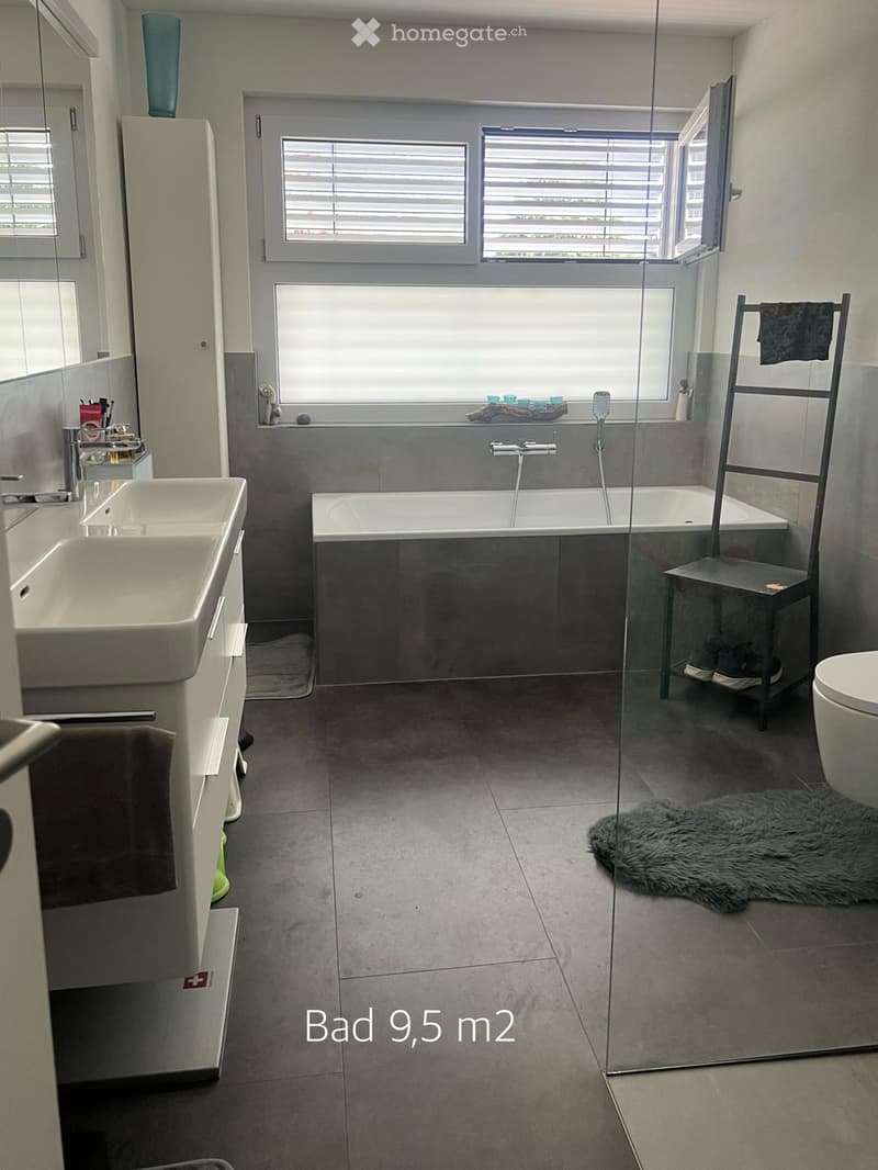 Modern bathroom with a bathtub, sink, toilet, mirror, and a towel rack. Features a large window with blinds and a fur rug on the floor.
