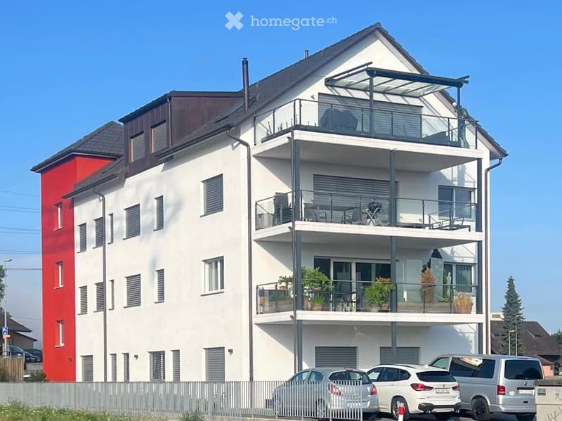 Modern two-story house with white and red facade, multiple balconies with glass railings, potted plants, and outdoor furniture.