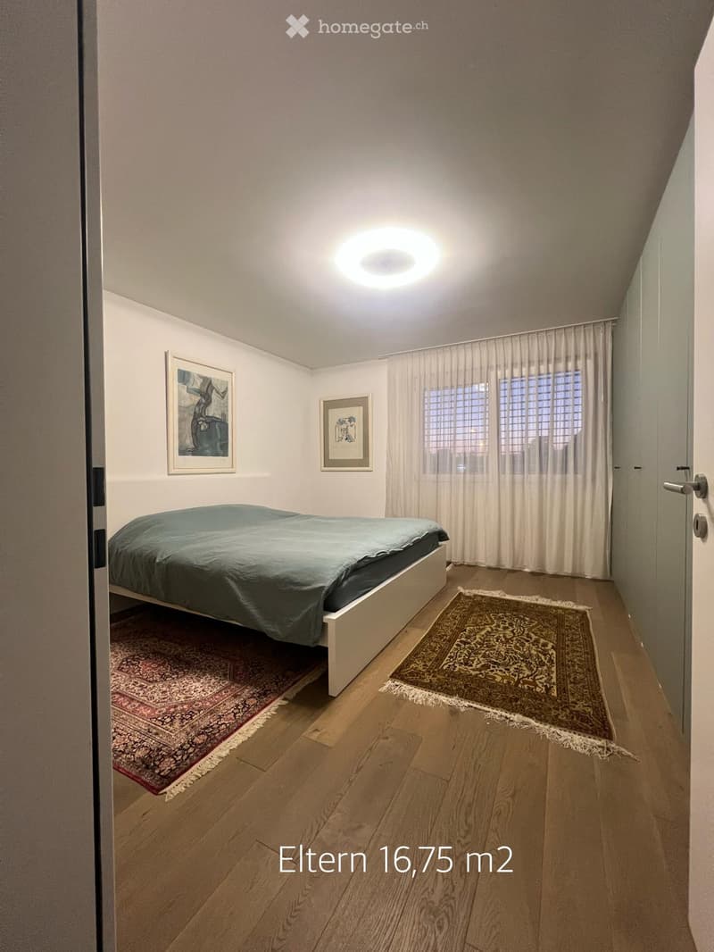 Modern bedroom with wooden flooring, a bed with a green duvet, two rugs, and two framed artworks on the wall.
