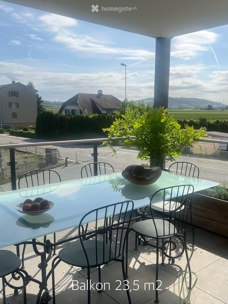 Balcony with a glass table, metal chairs, and a bowl of fruit, overlooking a scenic countryside view.