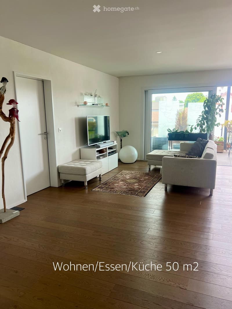 Modern living room with hardwood floors, large window, grey sofa, ottoman, coffee table, TV, decorative ball, and a unique wooden sculpture.