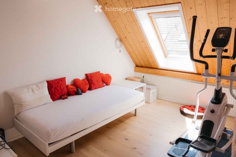 Cozy attic bedroom with a white bed, red heart-shaped pillows, a small table, and an exercise bike. The room features wooden slanted ceilings and large skylight windows.