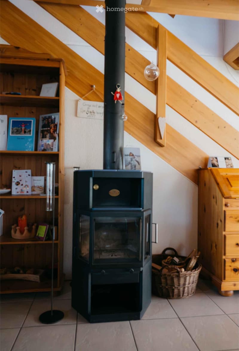 A cozy room with a modern black wood-burning stove, a wooden bookshelf with picture frames and books, and a basket of firewood nearby.