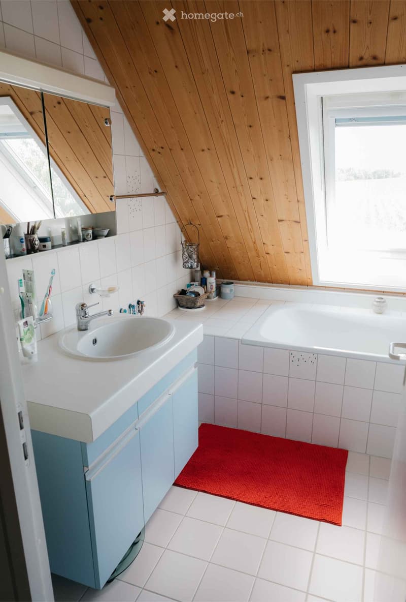 Bathroom with a white sink, bathtub, and wooden slanted ceiling. Includes a red bath mat, toothbrush holder, and small basket on the counter.