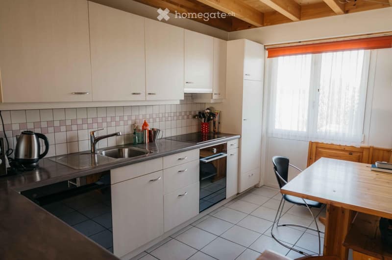 Modern kitchen with white cabinets, stainless steel sink, oven, and countertop. Includes a wooden dining table with chairs, kettle, and various kitchen items on the counter.