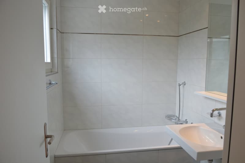 Modern bathroom with white tiles, a bathtub, a sink, a mirror, and a shelf.