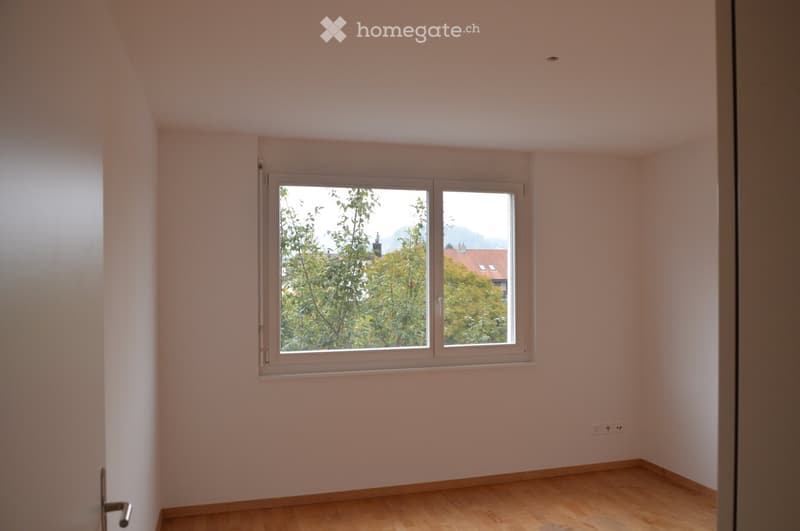 Empty room with light-colored walls, wooden flooring, and a large window offering a view of trees and a distant building.