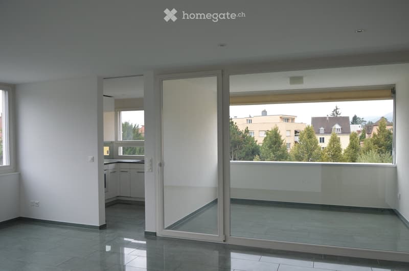 Empty room with tiled floor, white walls, and large glass sliding doors leading to a balcony with a view of trees and buildings.