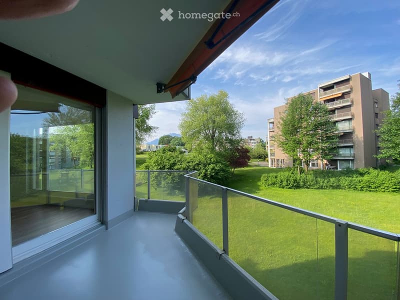 Balcony with glass railing, overlooking a green lawn and neighboring buildings.