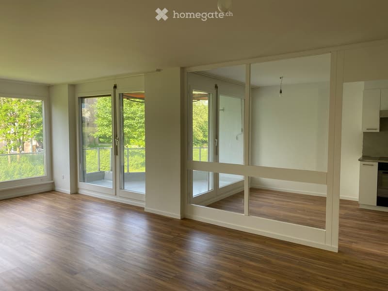 Empty room with wooden flooring, large windows, and sliding glass doors leading to a balcony area.