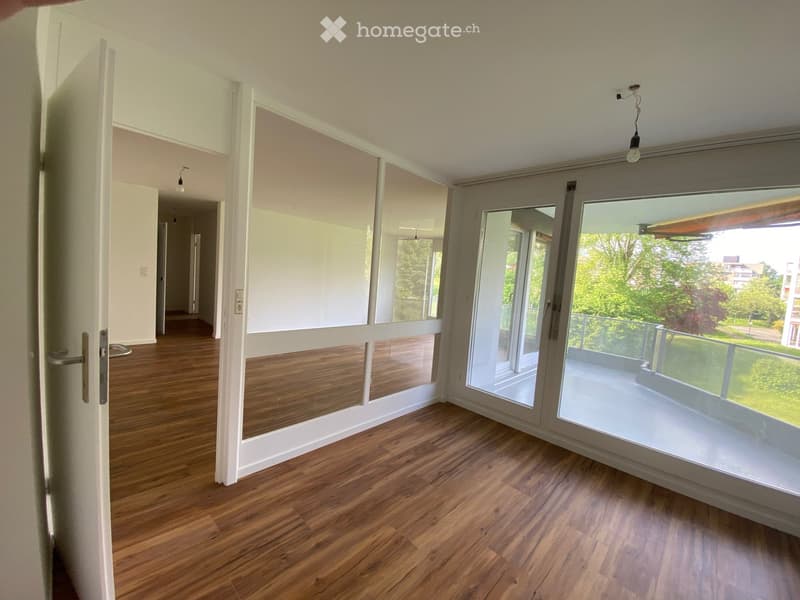 Empty room with wooden flooring, large windows, and sliding glass doors leading to a balcony. The space is well-lit with natural light and has a neutral color scheme.