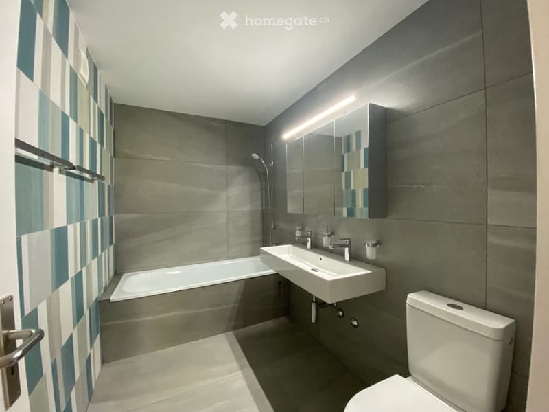 Modern bathroom with a bathtub, double sink, toilet, and a large mirror with built-in lighting. The walls are tiled in a grey and blue checkered pattern, and the floor is light grey.