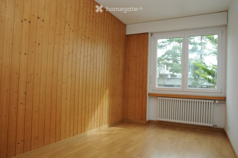Empty room with wooden walls and floor, large window with white frame, radiator below window, and white baseboard trim.