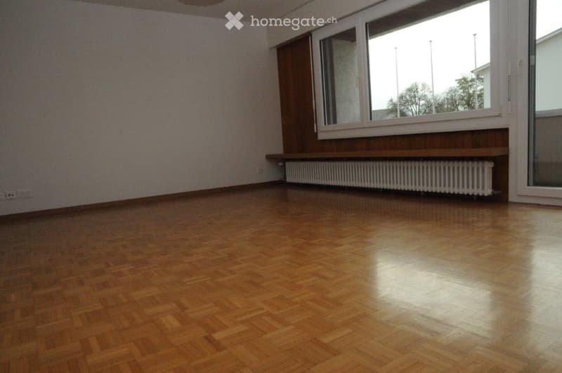 Empty room with wooden flooring, large windows, radiator, and patterned wallpaper.