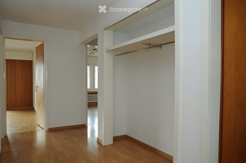 Empty hallway with wooden flooring, white walls, and built-in closet with shelves and a clothes rail. A wooden door is visible on the left, and a glimpse of another room can be seen through an open doorway.