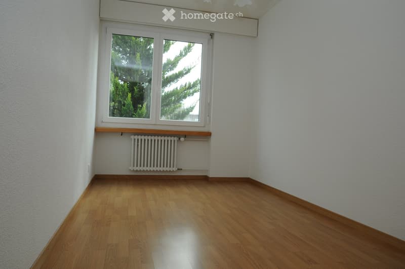 Empty room with wooden flooring, white walls, a window with a radiator below, and a small wooden shelf above the radiator.