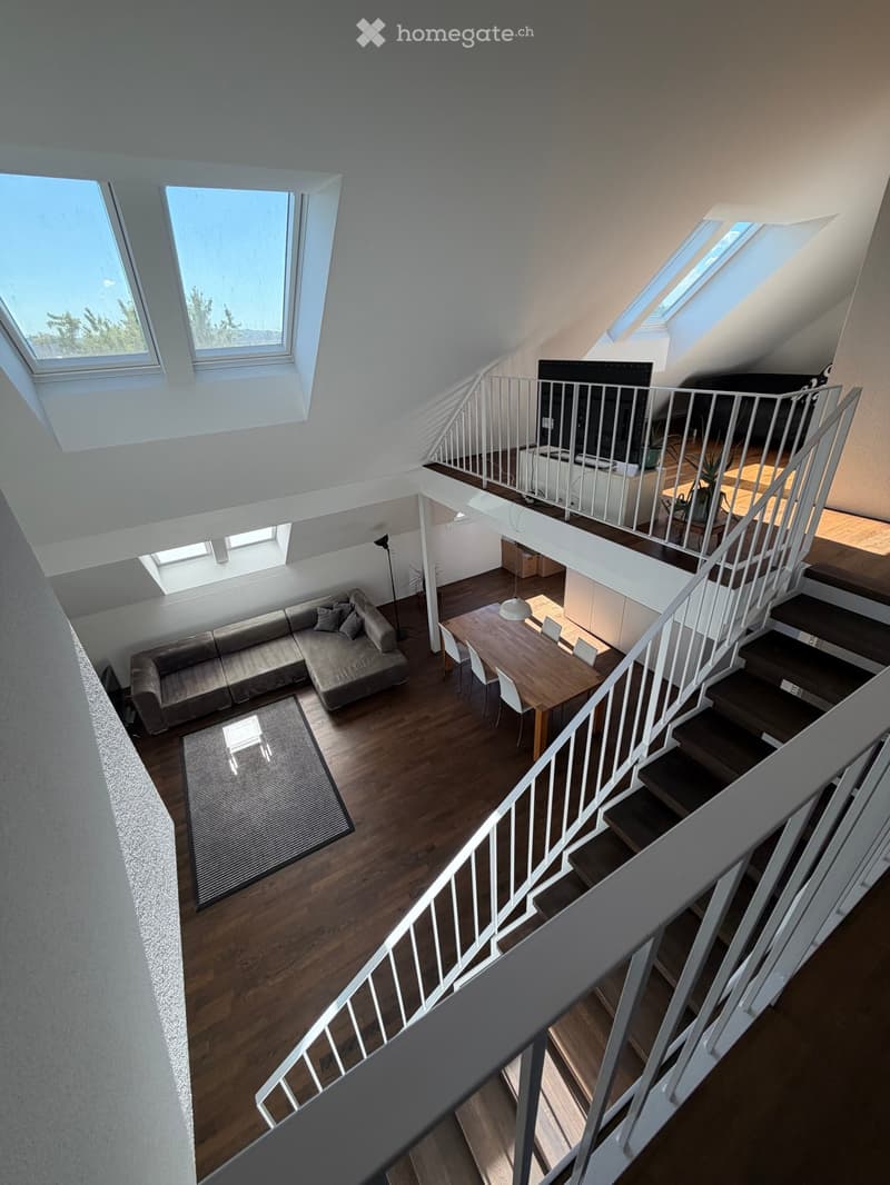 Open-plan living and dining area with wooden flooring, a sofa, dining table, chairs, and a staircase leading to an upper level with a TV screen.