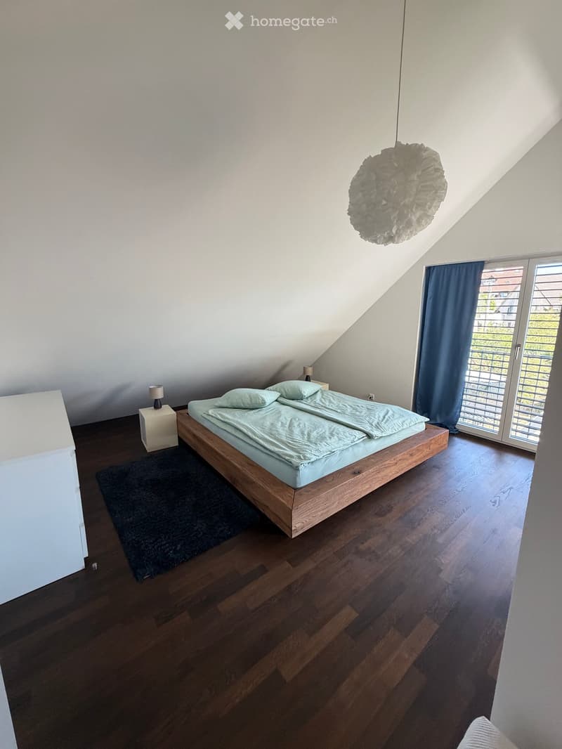 Modern bedroom with wooden floor, slanted ceiling, bed with light blue bedding, nightstand with lamp, and a large window with blue curtains.