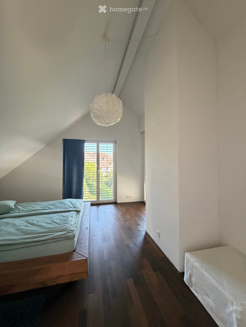 Bright attic bedroom with wooden flooring, two single beds, a large window with blue curtains, and a modern pendant light fixture.