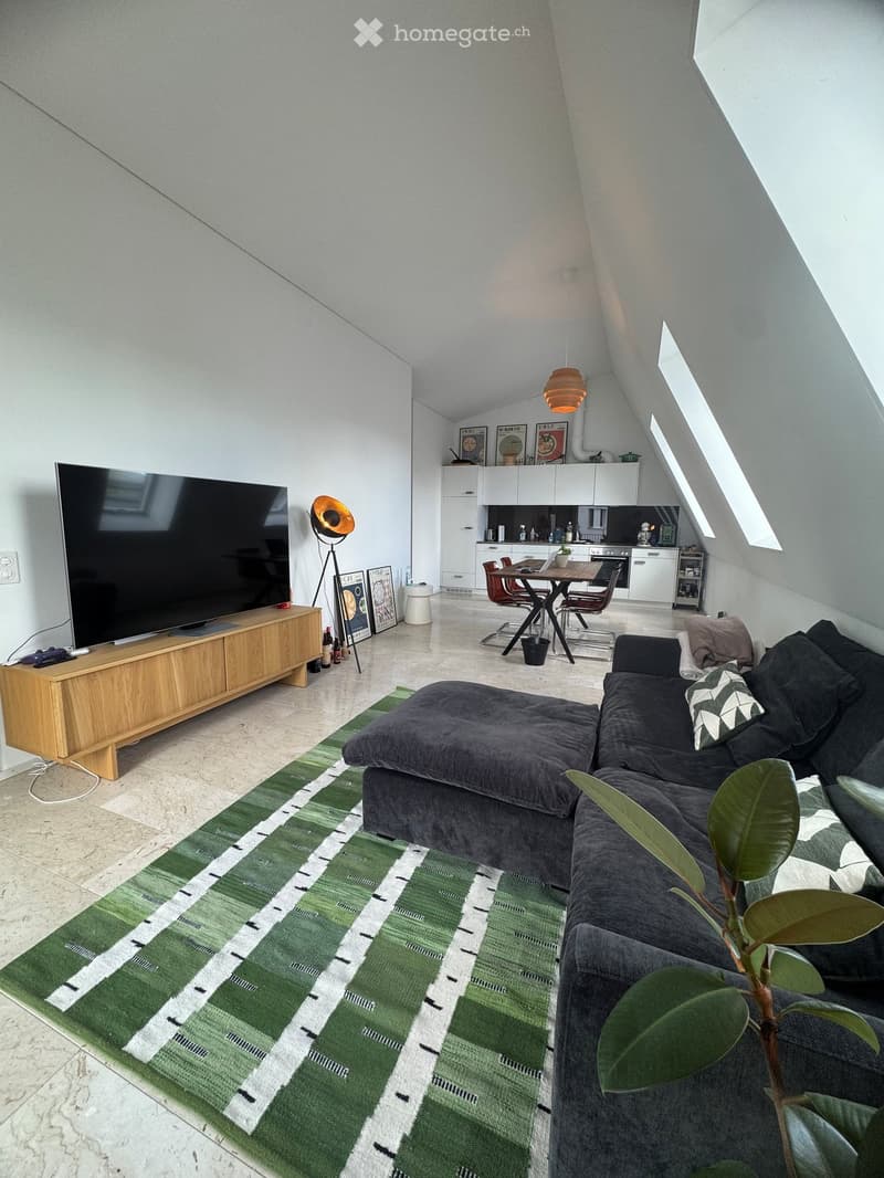 Modern living room with a grey sectional sofa, ottoman, coffee table, large television, floor lamp, dining area with table and chairs, kitchen area with cabinets, and a green patterned rug.