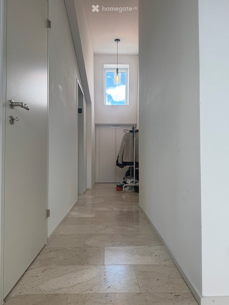 Modern hallway with light-colored walls, polished concrete floor, and a single window at the end. A coat rack with jackets and shoes is visible in the background.