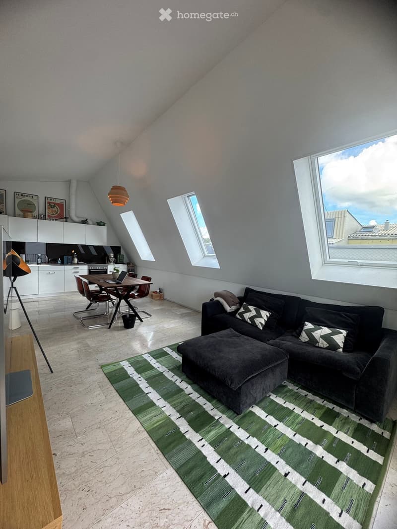 Bright, open living space with a sectional sofa, ottoman, coffee table, dining area with chairs and laptop, modern kitchen with white cabinets, and large windows letting in natural light.