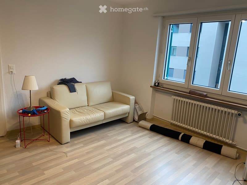 Living room with a light-colored sofa, a small red side table with a lamp, and large windows letting in natural light.